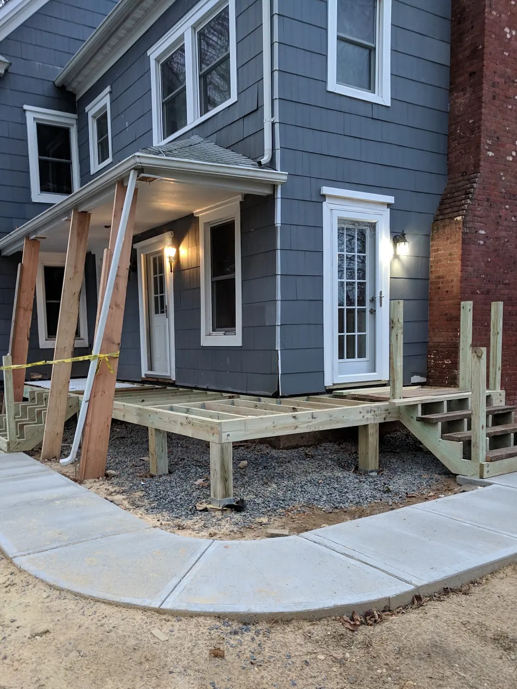 A front porch being framed in Monmouth County, NJ by RSC Contracting LLC: pressure-treated posts on metal bases over a gravel bed carrying the joists, the porch roof temporarily braced above, and a new concrete walk in front