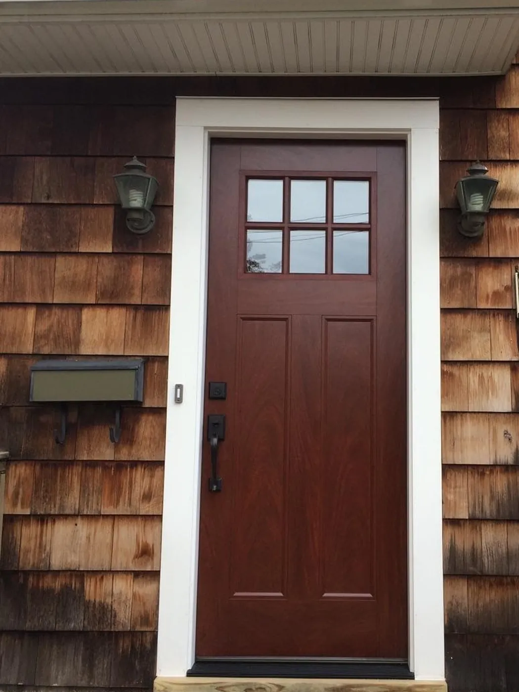 After: a new mahogany-stained craftsman entry door with a six-lite window and black hardware, cased in white trim, on the same cedar shake house