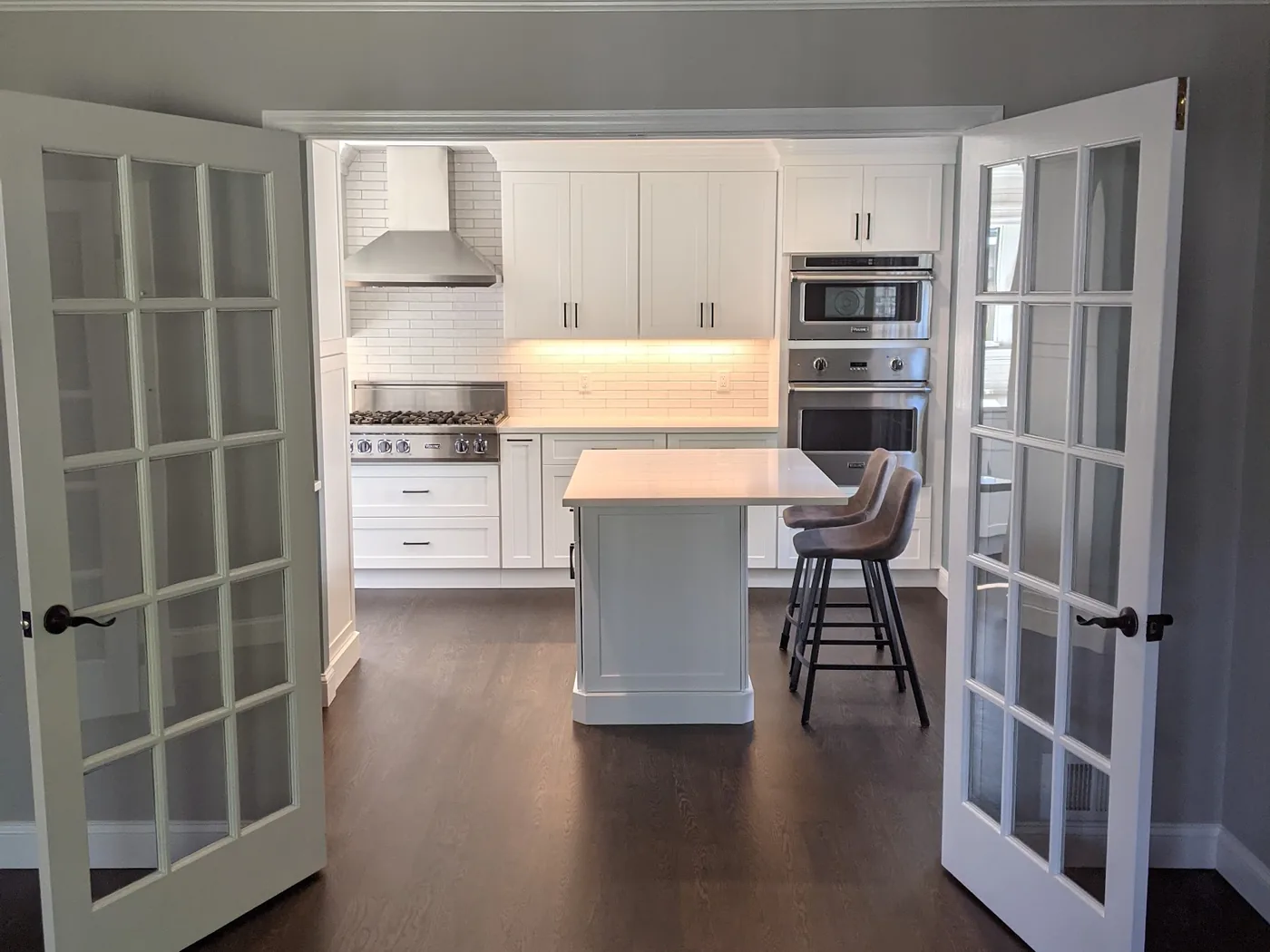 Opened-up first floor in Monmouth County, NJ seen through new French doors into a white kitchen with a seated island and stacked wall ovens